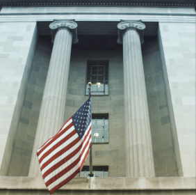 The American flag hangs between the grand columns of a neoclassical government building, likely the U.S. Department of Justice, symbolizing the federal agency's authority over the high-profile crypto fraud case mentioned in the post.