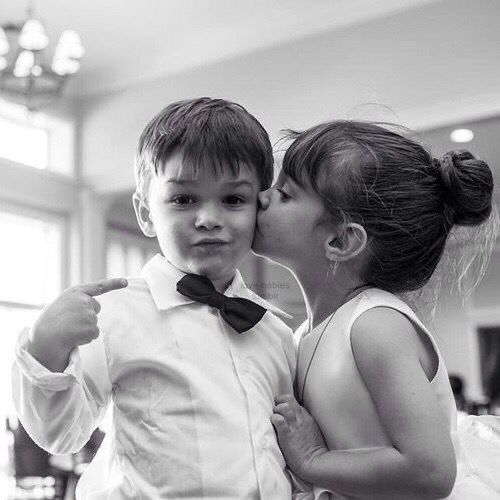 A black-and-white photograph shows a young boy in a bow tie smiling while a girl in a white dress kisses his cheek.