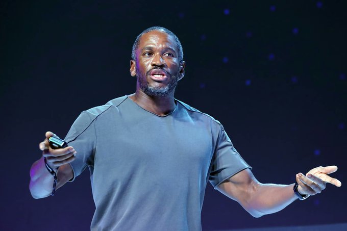 A man in a gray t-shirt speaks on stage with a microphone headset and a remote clicker in hand, set against a dark backdrop with starry lights. The image complements the post's theme, depicting what appears to be Arthur Hayes delivering a keynote on Bitcoin's need for monetary liquidity to break out of its current stagnation.