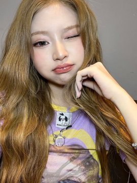 A close-up selfie of a young woman with long blonde hair, pink blush, and glossy lips wearing a purple tie-dye shirt with a small pendant, posing with her hand near her face against a grey wall.