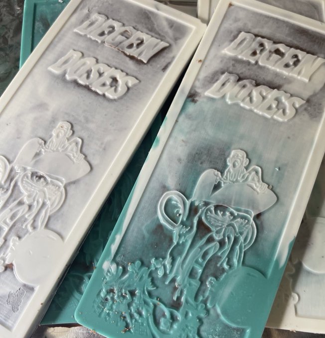 Two freshly mixed resin bars lie side by side, each featuring raised lettering that reads "DEGEN DOSES" above a 3D molded design of a pill-bottle-shaped figure with mushroom imagery, coated in white and teal pigments that give them a marbled, frosted appearance—capturing the early curing stage of @degendoses' bar-making process.