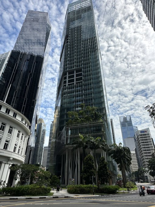 A motorcyclist rides past towering glass skyscrapers and palm trees in Singapore's Marina Bay area, with the DBS building visible in the distance. The urban backdrop sets the scene for a busy day of crypto side events around Marina Bay Sands and beyond.