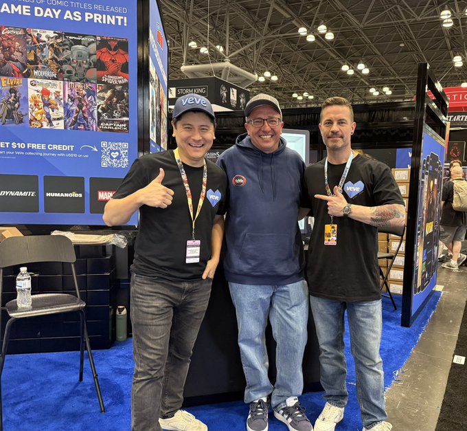 Three men smiling at a comic convention booth for 'veve' displaying 'COMIC DAY AS PRINT!' signage with titles like Spider-Man and Captain America visible.