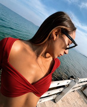 A young woman with long dark hair wearing a red crop top and sunglasses, taking a selfie outdoors near water with a pier visible in the background.