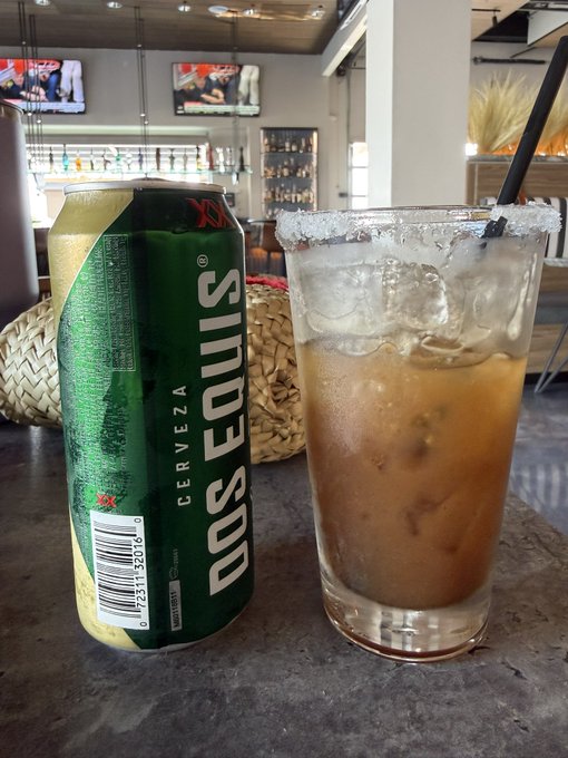 A can of Dos Equis beer sits next to a salt-rimmed mixed drink on a bar counter, with a bar area and TV screens visible in the background.