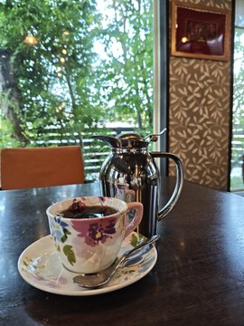 A floral teacup and saucer sit on a dark wooden table beside a glass teapot, with a window showing green trees in the background.