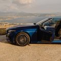 A small image of a dark blue convertible sports car parked on a gravel road overlooking a scenic landscape with hills in the background.