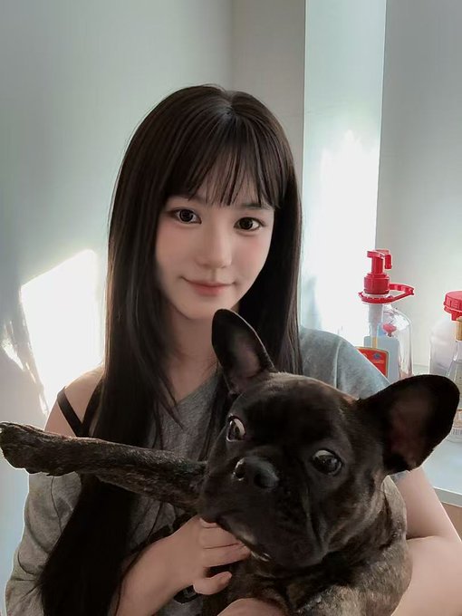 A young woman with long dark hair holds a French Bulldog in front of a window with cleaning supplies visible in the background.