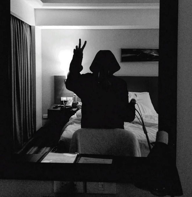 A black-and-white photo showing a silhouetted figure sitting on a hotel bed in front of a mirror, holding up a peace sign.