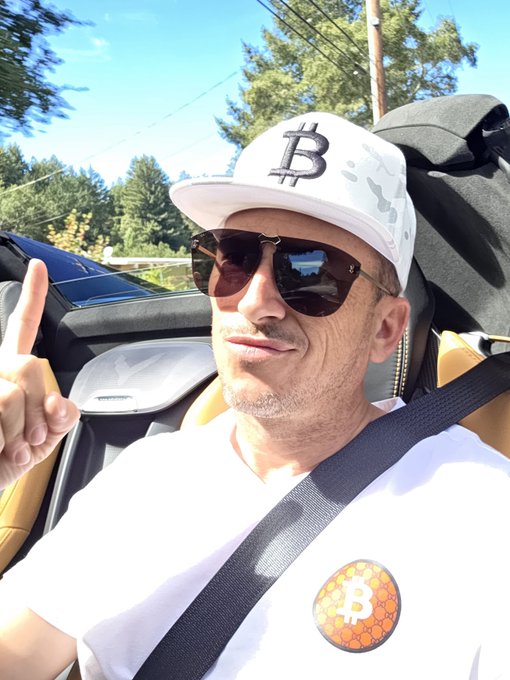 A man sits in a convertible car wearing sunglasses, a white t-shirt with a Bitcoin logo patch, and a white cap featuring a black Bitcoin symbol, giving a peace sign.