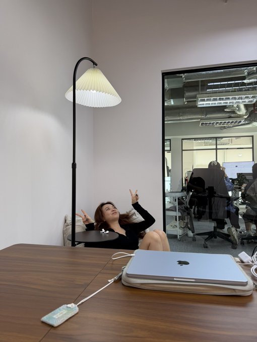 A woman sits casually in a chair next to a tall floor lamp and a small round table in a modern office space, gesturing with both hands. In the foreground, a silver laptop and a connected phone rest on a wooden table.