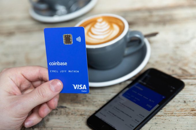 A hand holding a blue Coinbase Visa debit card (personalized 'CYRIL JEFF MATHEW') next to a smartphone and a cup of coffee.