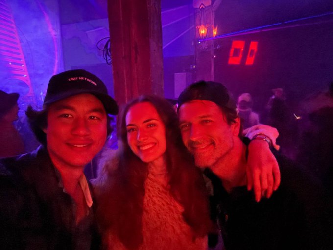 A selfie of three people smiling in what appears to be a club or bar with red and purple lighting, with one person wearing a black cap with red text.