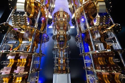 A close-up of an IBM Quantum System One or similar quantum computer, showing its ornate gold chandelier-like dilution refrigerator structure illuminated with blue light.