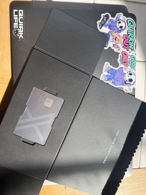 A black shipping box with 'QUIRK LIFE' branding and a colorful sticker reading 'QUIRK ONLY VESA', opened to reveal a compartment labeled 'TAP INTO EVERYTHING' with a SIM card holder.