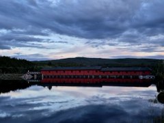 A serene landscape at twilight shows a red train traveling across a bridge reflected in still water, with cloudy skies above.