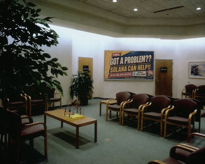 A waiting room with rows of burgundy chairs faces a wall banner that reads 'Folks GOT A PROBLEM?? SOLANA CAN HELP!!' listing contact information for the Solana Network.