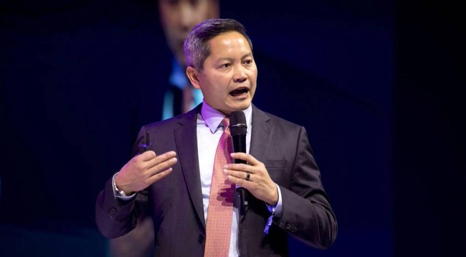 A man in a dark suit with a salmon-colored tie holding a microphone while speaking at what appears to be a conference or presentation.