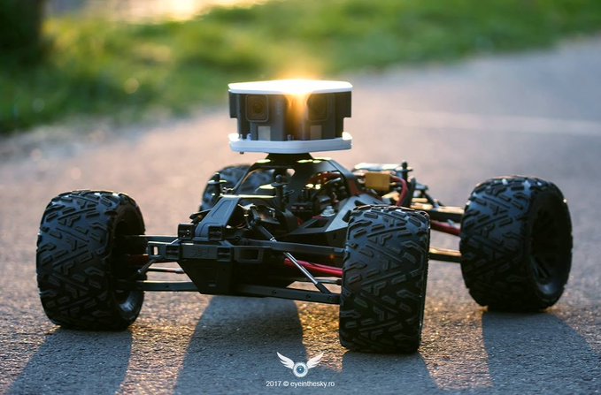 A small autonomous robotic car with four rugged tires and a top-mounted LiDAR sensor parked on pavement in golden sunlight, watermarked '2017 © eyenteksy.ro'.