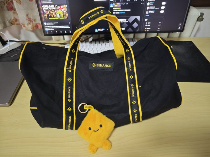 A black and yellow Binance-branded duffel bag with "BINANCE" text on the straps and a "BINANCE" logo patch sits on a wooden desk, accompanied by a small orange BIBI plush keychain hanging from the bag's handle. The merchandise in the image matches the post celebrating the arrival of Binance's latest swag, a travel bag and the BIBI charm.