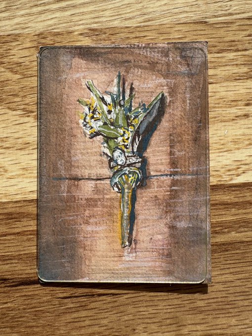 A small painting or mixed-media artwork depicts a green, leafy bouquet or floral arrangement against a brown background, resting on a wooden surface.