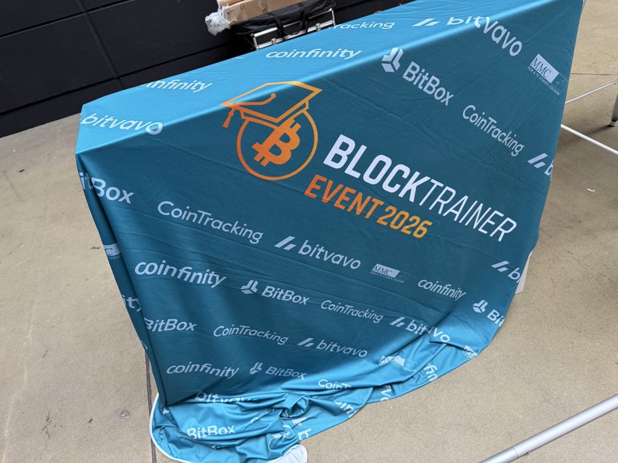 A blue event banner being set up, featuring the 'BLOCKTRAINER EVENT 2026' logo with a Bitcoin graduation cap icon, surrounded by sponsor logos including BitBox, Coinfinity, Bitvavo, CoinTracking, and MIC.
