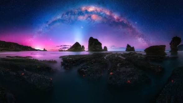 A stunning nightscape featuring a colorful Milky Way arch over a rocky coastline with sea stacks and a reflective tide pool illuminated by pink and purple hues.