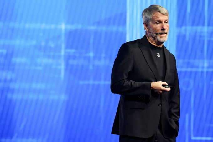 Michael Saylor, wearing a black suit with a Bitcoin logo pin, speaking on stage with a blue digital background.
