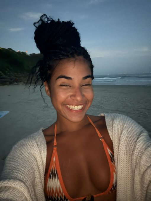 A smiling woman with her hair in a high bun stands on a beach at dusk, wearing an orange patterned bikini top and a cream-colored knit cardigan.