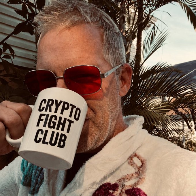 A man wearing red-tinted sunglasses and a white robe drinks from a white mug printed with 'CRYPTO FIGHT CLUB' in bold black letters, photographed outdoors with palm fronds in the background.