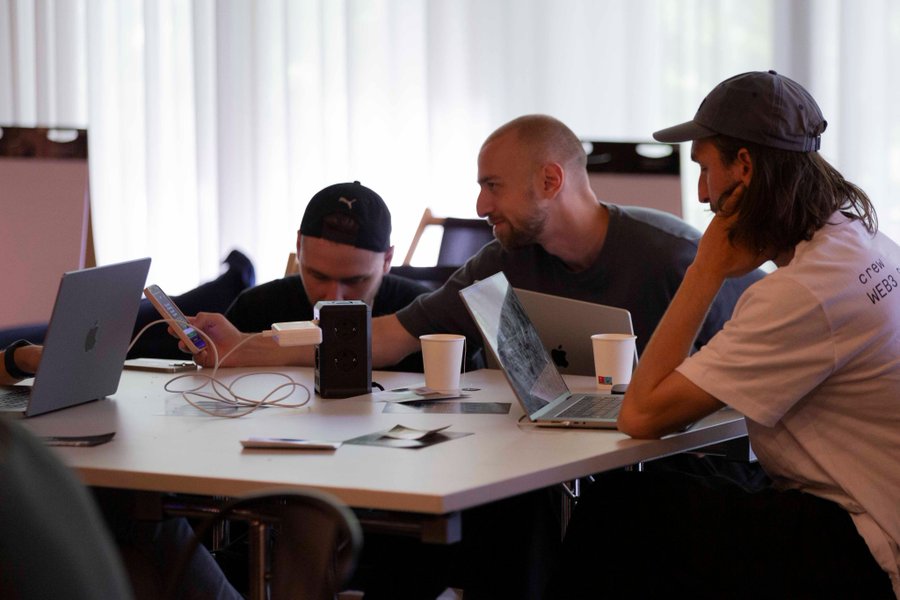 Three developers huddle around laptops and coffee cups at a cluttered table, with one wearing a shirt partially reading "CTDN WEB3," capturing the collaborative, first-time shipping atmosphere described in the post about 190 developers and 272 .dot domains at Funkhaus.