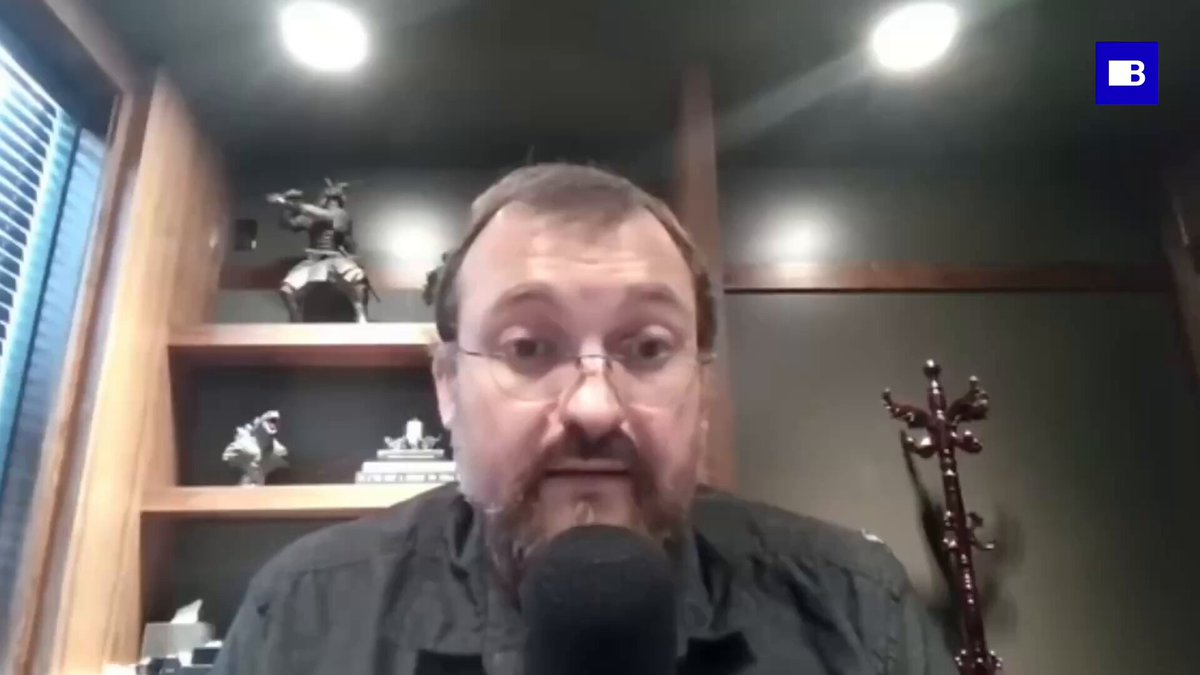 A man wearing glasses and a black shirt with a microphone in front of him, which could be used for a podcast or video recording session. He is seated in front of a bookshelf filled with various objects including sculptures and vases that are arranged on different shelves. The image suggests an atmosphere where Charles Hoskinson is sharing his thoughts, possibly discussing the value of money or comparing it to something humorous about toilet prices.