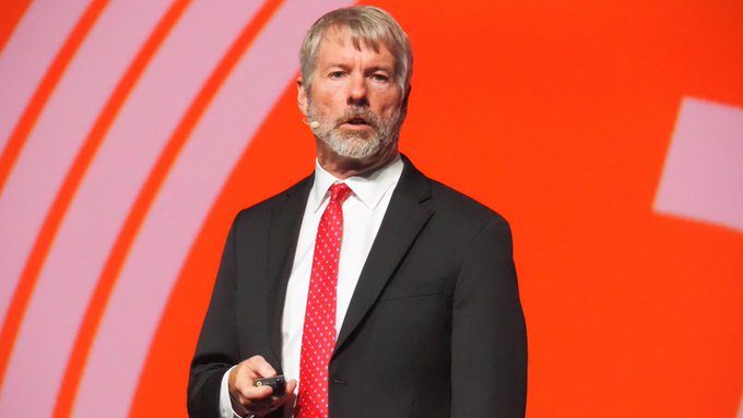 A man wearing a suit and tie standing in front of an orange background with text that says "MICHAEL SAYLOR'S BITCOIN STRATEGY IS NOW DOWN $11.5 BILLION IN UNREALIZED LOSSES."