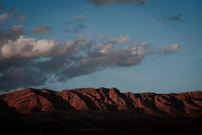 A scenic landscape showing rugged red rock formations bathed in warm light, set against a deep blue sky scattered with soft white clouds at dusk.