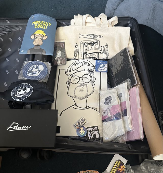 An open suitcase filled with Bored Ape Yacht Club (BAYC) merchandise, including a 'Mindfully Bored' book, a canvas tote bag, patches, a black fanny pack, an artist-signed print of the NFT collection, and a PUMA shoebox.