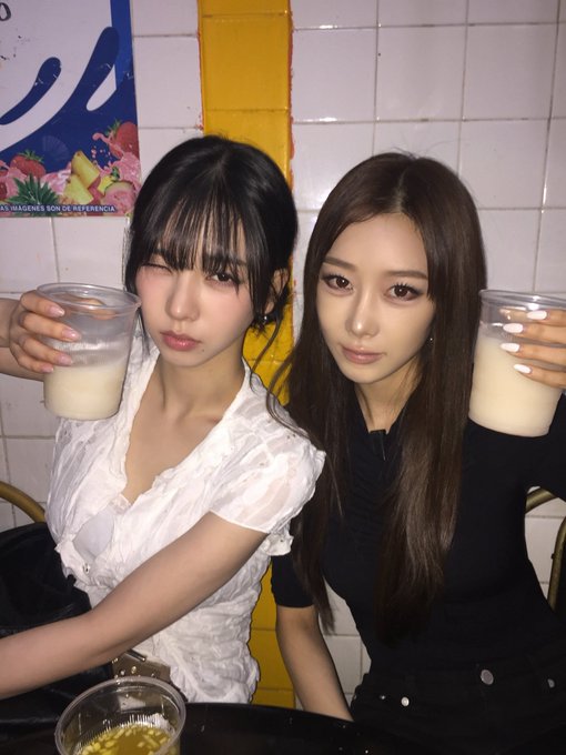Two young women posing together at a table, each holding up a clear cup of milk, with a yellow tiled wall and tropical drink poster in the background.