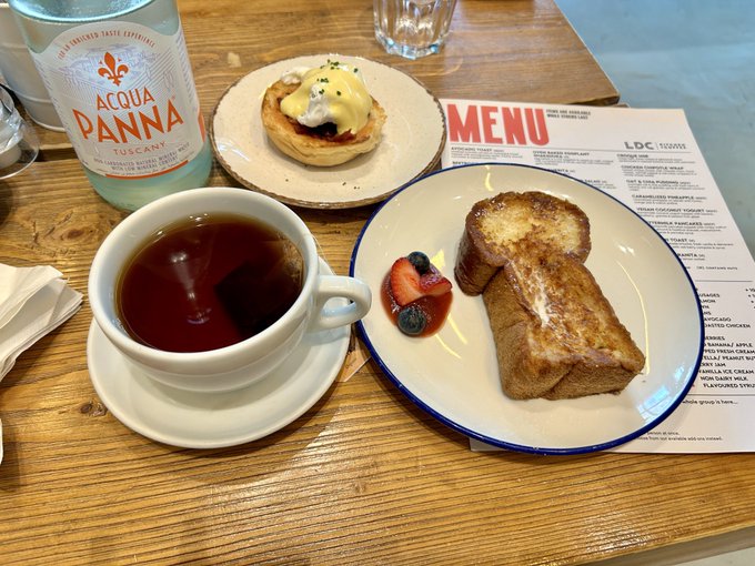 A brunch spread featuring Acqua Panna water, a cup of tea, an eggs benedict dish, and French toast with berries and cream on a wooden table next to a menu.