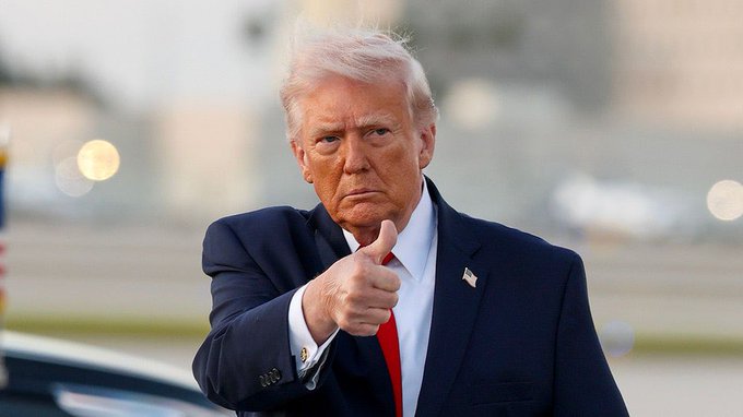 Donald Trump, dressed in a dark suit and red tie, gives a thumbs-up gesture with a serious expression while standing outdoors.