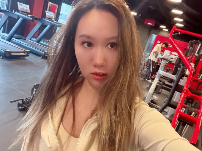 The image features a woman with long hair, wearing a white shirt, posing for a photo at a gym or fitness center. The background of the picture shows various exercise equipment such as treadmills and weight machines. There are also two people present in the scene, one standing near the left side of the image and another person closer to the right side.