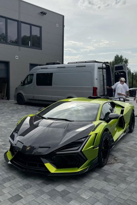 A bright green Lamborghini Revuelto sports car parked on a driveway in front of a gray Sprinter-style van and a modern building, with a person standing near the van.