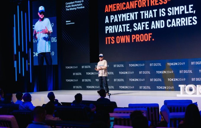 A presenter on stage at the TOKEN2049 conference with a large screen displaying 'AMERICANFORTRESS™: A PAYMENT THAT IS SIMPLE, PRIVATE, SAFE AND CARRIES ITS OWN PROOF.'
