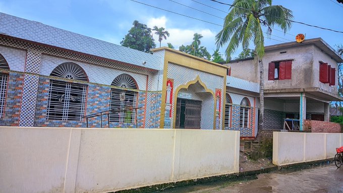 A street-level view of colorful houses in what appears to be a tropical residential area, with checkered tile walls, a satellite dish, and palm trees visible.