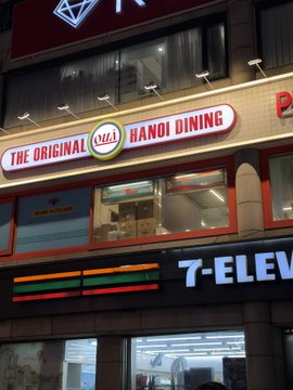 An illuminated sign for 'THE ORIGINAL VIET HANOI DINING' is mounted above a 7-Eleven storefront at night.