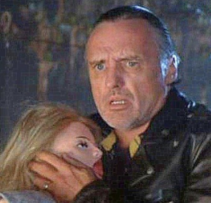 A man wearing a yarmulke grabs a blonde woman by the face while both appear startled, seemingly in a scene from an action or thriller film. The accompanying post contains only a random alphanumeric string with no discernible connection to the image.