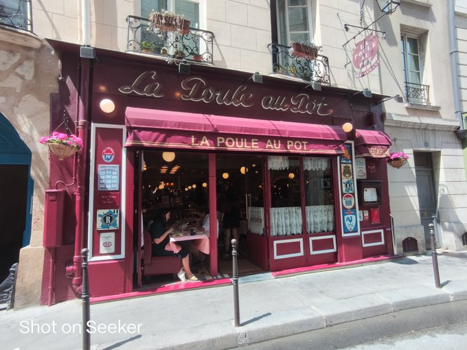A photograph of a red-fronted French restaurant named 'La Poule au Pot' / 'la Douce au Pot' with awnings, flower boxes, and patrons dining inside, captioned 'Shot on Seeker'.