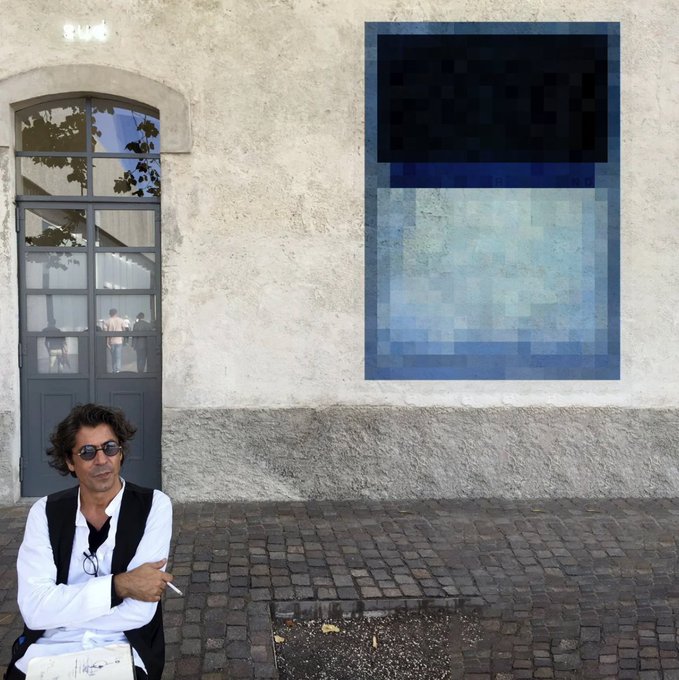A man with sunglasses sits on a cobblestone street in front of a weathered wall displaying a blue and black abstract painting.
