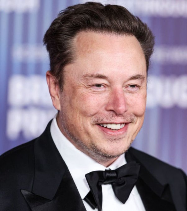 A photograph of Elon Musk smiling in a tuxedo at what appears to be a formal event.