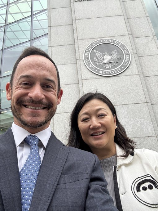 A selfie of a smiling man in a gray suit and a smiling woman in a cream jacket taken in front of the U.S. Securities and Exchange Commission (SEC) building seal.