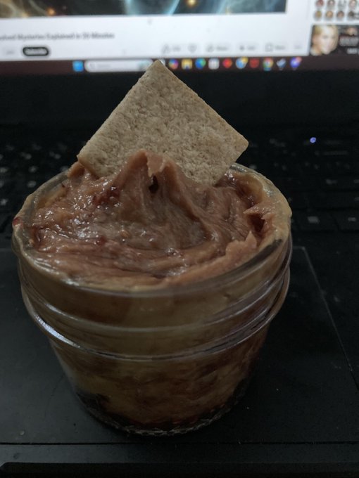The image features a jar of peanut butter with a square piece of bread sticking out of it. The jar is placed on top of a black surface, possibly a desk or table. The scene appears to be set up for a photo shoot or an artistic composition that highlights the contrast between the peanut butter and the bread.