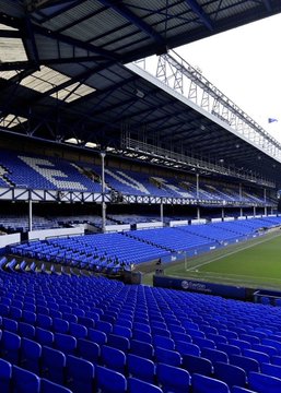 An empty blue-seated stadium stand at Goodison Park, Everton's home ground, awaiting the special charity match where the winning VIP bidder will experience a day in the life of an Everton player. The pristine seating area highlights the exclusive nature of the experience that includes hotel stays with the team, training sessions, and a bench spot during the game.